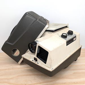May include: A vintage white and gray slide projector with a black lens and a gray carrying case. The projector has a label that reads "Kodak Carousel 805".