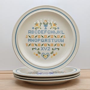 Könnte beinhalten: Ein Stapel cremefarbener Keramikteller mit blauem Rand und einem Kreuzstich-Alphabet-Design. Die Teller haben einen hellblauen Rand und ein dekoratives Muster in Gelb, Grün und Blau. Die Teller sind auf einer Holzoberfläche gestapelt.