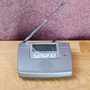 May include: A silver RadioShack weather radio with a digital display showing 12:04 PM and 12:00, a central speaker grille, and a long, thin, extendable metal antenna. The device features buttons labeled Weather and Snooze below the screen.