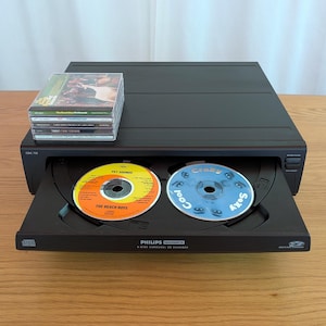 Może przedstawiać: Czarny zmieniacz płyt CD Philips Magnavox z 6 płytami w karuzeli z dwoma płytami CD widocznymi w tacy. Jedna płyta CD jest oznaczona jako "Pet Sounds" The Beach Boys. Na górze znajduje się stos płyt CD. Tekst "Cool" i "Sexy" jest widoczny na drugiej płycie CD.