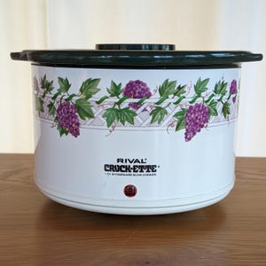以下が含まれることがあります： ダークグリーンの蓋と縁が付いた、白いRival Crock-Ette 1Lストーンウェアスロークッカー。スロークッカーには、紫色のブドウと緑の葉の装飾バンドが付いています。「RIVAL CROCK-ETTE」の文字が前面に印刷されています。