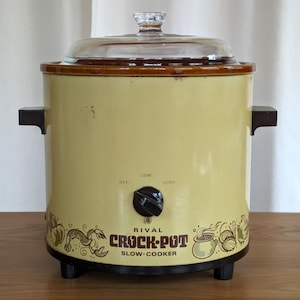 May include: Vintage Rival Crock-Pot slow cooker in a pale yellow color. It has a glass lid and brown trim. The control knob has settings for off, low, and high. The Crock-Pot has black handles and feet, and the text "RIVAL CROCK-POT SLOW-COOKER" is printed on the front.