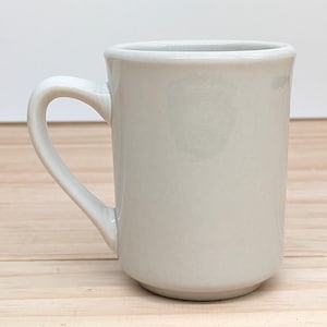 Peut inclure: Une tasse en céramique blanche unie avec une anse. La tasse est vide et a une finition lisse et brillante.