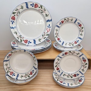 May include: A collection of white ceramic plates, bowls, and saucers with a blue scalloped edge and floral design. The plates feature red and green floral accents and blue decorative elements. The set includes various sizes of plates and bowls.