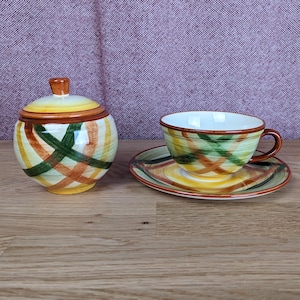 May include: A ceramic teacup, saucer, and sugar bowl set. The set features a colorful plaid pattern in shades of green, yellow, orange, and brown. The teacup has a handle and sits on a matching saucer. The sugar bowl has a lid.
