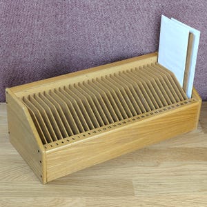 May include: A light-colored wooden desk organizer featuring thirty-one vertical slots for document storage. Each slot is numbered sequentially from 1 to 31 in black ink along the front edge. A white envelope is placed in the final slot on the right.