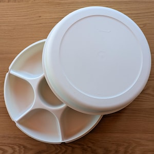 Puede incluir: Bandeja de servir de plástico blanco con tapa. La bandeja tiene cinco secciones para servir diferentes alimentos. La tapa es lisa y tiene la palabra "TUPPERWARE" impresa.