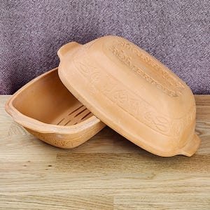 May include: A rectangular terracotta Römertopf baking dish with a matching lid. The lid features embossed leaf and bird motifs along the edges and the text Römertopf. The base has a slotted bottom and integrated handles on both sides.