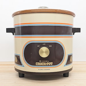 Peut inclure: Un autocuiseur Rival Crock-Pot vintage de couleur crème, marron et orange. L'autocuiseur a un couvercle et des poignées marron, avec un bouton de commande étiqueté "Off", "Low" et "High". Le texte "RIVAL CROCK-POT SLOW-COOKER" est affiché à l'avant.