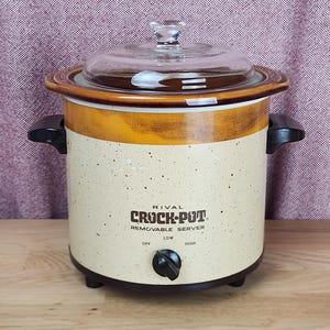 Könnte beinhalten: Ein Vintage Rival Crock-Pot Slow Cooker mit gesprenkeltem beigefarbenem Äußeren, braunen und goldenen Akzenten und einem Deckel aus klarem Glas. Der Bedienknopf ist schwarz, und der Text "Crock-Pot" ist auf der Vorderseite sichtbar. Der Slow Cooker hat schwarze Griffe.