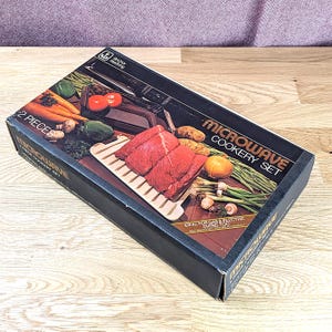 Könnte beinhalten: Ein schwarzer rechteckiger Karton für ein Anchor Hocking Mikrowellen-Kochset. Die Verpackung zeigt eine Grafik eines zweiteiligen Bratgestells und einer Schale mit rohem Rindfleisch, Karotten, Kartoffeln und grünen Bohnen.