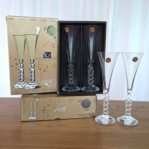 Puede incluir: Varias copas de champán con cuencos transparentes en forma de cono y tallos decorativos se exhiben. Algunas están en cajas con el texto "Millennium" y "Cristal d'Arques". Las copas tienen un diseño único, con el número "2000" en el tallo.