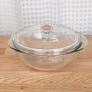 May include: A clear glass casserole dish with a matching lid. The dish has a rounded shape with two handles and a clear glass lid with a decorative knob. The dish is sitting on a wooden surface.