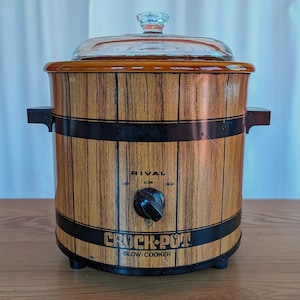 May include: Vintage Rival Crock-Pot slow cooker with a wood-grain design. The appliance features a clear glass lid with a knob, a black control knob, and the text "RIVAL" and "CROCK-POT SLOW-COOKER". The slow cooker has black bands around the middle and bottom.
