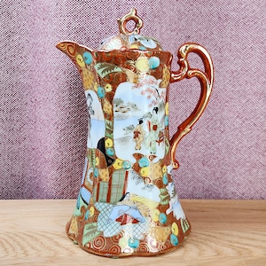 May include: A ceramic coffee pot featuring a glossy finish with intricate orange, gold, and blue patterns. The body displays detailed illustrations of figures in traditional attire within landscape scenes, complemented by a curved handle and lid.