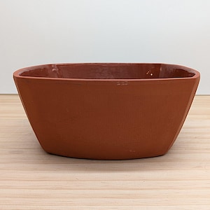 May include: A terracotta-coloured, rectangular bowl with a slightly tapered design. The bowl has a smooth exterior and a darker, textured interior. The bowl is sitting on a light-coloured wooden surface.