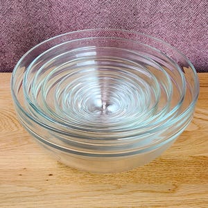 May include: A set of five clear glass mixing bowls nested inside one another. Each bowl features a smooth, transparent surface with a rounded base and a wide rim. The bowls are stacked in descending order of size, creating a concentric circular pattern.