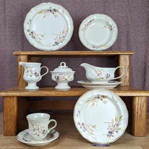 May include: A vintage cream-coloured dinner service featuring floral patterns. The set includes plates, bowls, a creamer jug, a sugar bowl, a gravy boat, and a teacup with saucer. The edges of the items have a scalloped design and gold trim.