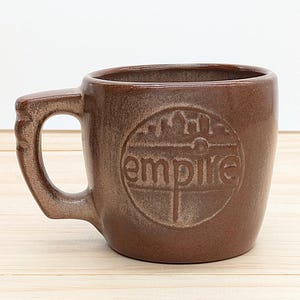Puede incluir: Una taza de cerámica marrón con asa. La taza presenta la palabra "empire" en un diseño circular. La taza es de color marrón cálido y parece estar hecha de gres. La taza está sobre una superficie de madera de color claro.