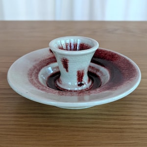 Puede incluir: Un portavelas de cerámica con base color crema y diseño de círculos concéntricos rojo oscuro. El portavelas presenta una copa central acampanada para la vela y un plato circundante. La pieza tiene un acabado de esmalte craquelado, lo que añade a su estética vintage.