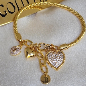 Puede incluir: Pulsera dorada con un diseño de cuerda trenzada y varios colgantes. Los colgantes incluyen un corazón, un candado y un círculo con la letra 'B' en él. Todos los colgantes están adornados con brillantes diamantes de imitación transparentes.