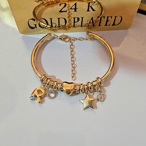 Puede incluir: Un brazalete chapado en oro con un estilo de brazalete de abalorios. El brazalete tiene un abalorio en forma de corazón que dice "love", un abalorio en forma de estrella, un abalorio con un ojo turco azul y un abalorio con la letra "P". El brazalete está chapado en oro de 24 quilates.