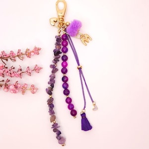 Puede incluir: Un llavero dorado con un cierre de mosquetón, adornado con cuentas moradas, una pequeña borla morada y un acento morado esponjoso. El llavero también presenta un imperdible dorado y las letras "ME".