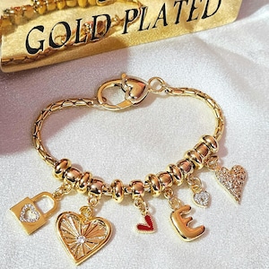 Puede incluir: Un brazalete de charms dorado con charms en forma de corazón, candado y letras. El brazalete está hecho de metal chapado en oro y tiene un cierre de langosta.