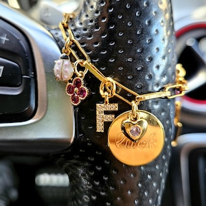 Op de afbeelding: Een goudkleurige schakelarmband met diverse bedels: een wit met roze lieveheersbeestje, een rood klavertjevier, een letter F bezet met heldere steentjes en een ronde gouden schijf met de inscriptie luck en een klein hartje met roze steen.