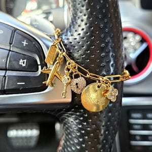 May include: A gold-toned chain bracelet featuring multiple charms including an airplane, a key, a heart-shaped padlock with embedded stones, a round disc engraved with the word "Luck", a four-leaf clover charm, and a rectangular tag labeled "LOVE".