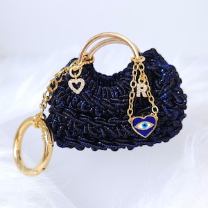 May include: A small, navy blue crocheted handbag with a gold handle and accents. The bag features a gold chain with a heart charm and a chain with a blue heart-shaped charm with an eye design. The bag is embellished with gold hardware.