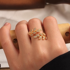 Op de afbeelding: Een goudkleurige ring met twee overlappende, bladvormige takken. Elk blaadje is gedetailleerd met kleine, heldere, fonkelende steentjes die op het oppervlak zijn gezet. De ring heeft een open ontwerp, waardoor de motieven om de vinger vallen.