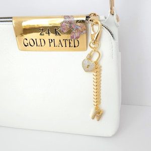 May include: A white handbag with a gold-plated accent featuring the text "24K GOLD PLATED" and a butterfly design. A gold charm with a heart-shaped lock, a fishbone design, and the letter "H" hangs from the zipper.