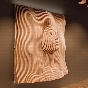 Op de afbeelding: Een houten wandsculptuur van een gezicht, gemaakt met gelaagde houten stroken. De sculptuur is gemaakt van lichtbruin hout en heeft een 3D-effect.