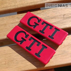 Op de afbeelding: Twee rode rechthoekige GTI-emblemen met zwarte letters worden weergegeven op een houten oppervlak. De emblemen zijn schuin geplaatst en de letters zijn vetgedrukt en gemakkelijk te lezen. De achtergrond is een lichtbruin houten oppervlak.