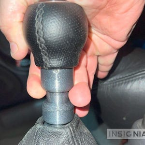 May include: A black leather gear shift knob with a gray base is held by a hand. The knob has a stitched seam and a textured surface. The base is cylindrical and connects to a black leather boot.
