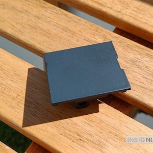 Puede incluir: Una cubierta rectangular de plástico negro mate con una superficie superior plana y una pequeña muesca en el lado derecho. El componente tiene una base central que sobresale por debajo, diseñada para su fijación, con el texto INSIGNIAS3D visible.