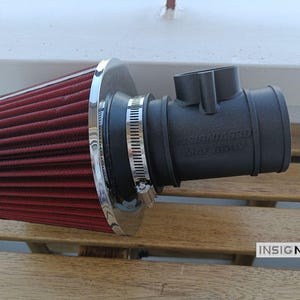 Może przedstawiać: Stożkowy filtr powietrza z ciemnoczerwonego plisowanego materiału z polerowaną chromowaną nasadką, przymocowany do matowego czarnego cylindrycznego adaptera wlotowego. Adapter posiada metalową opaskę zaciskową oraz wytłoczony napis INSIGNIAS3D MAF BODY.
