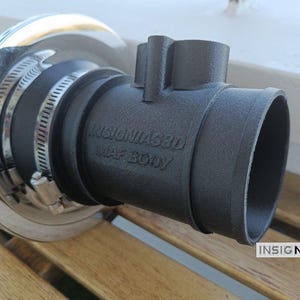 Może przedstawiać: Czarny matowy adapter korpusu MAF z wytłoczonym napisem INSIGNIAS3D MAF BODY, połączony z cylindrycznym czerwonym filtrem powietrza z chromowaną metalową krawędzią za pomocą dw&oacute;ch srebrnych metalowych opasek zaciskowych. Element ma teksturowane wykończenie.