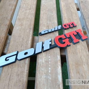 Peut inclure: Deux emblèmes Golf GTI imprimés en 3D, rouges, gris et noirs, posés sur une surface en lattes de bois. Le plus grand emblème est au premier plan, avec un plus petit au-dessus. Le mot "Golf" est en gris et "GTI" en rouge.