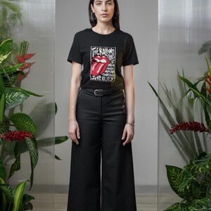 May include: Black t-shirt featuring the Rolling Stones' iconic tongue and lips logo with text. The shirt is paired with black pants and white sneakers. The image is set against a neutral background with plants.