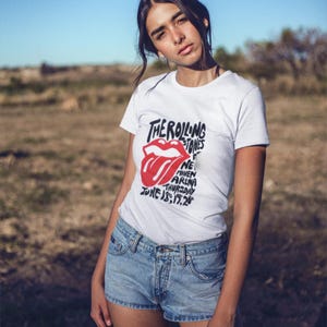 May include: White t-shirt featuring the Rolling Stones logo with a red tongue graphic and text. The shirt also includes the text "New Haven Arena Thursday June 18, 1972". The model is wearing denim shorts.