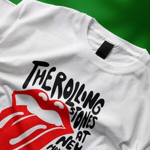May include: White t-shirt featuring the iconic Rolling Stones tongue and lips logo in red, with black text. The text reads "THE ROLLING STONES AT NEW HAVEN". The shirt is made of a soft, white fabric.
