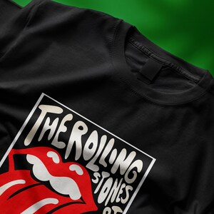May include: Black t-shirt featuring the iconic Rolling Stones tongue and lips logo. The design is in a white rectangular frame with the band's name in a stylized font. The red lips and tongue are a prominent feature.