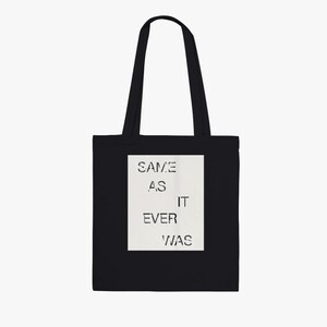 Puede incluir: Bolso tote negro con asas largas. Un rectángulo blanco en la parte delantera muestra el texto "SAME AS IT EVER WAS" en una fuente de plantilla. La bolsa está hecha de un material resistente y es adecuada para transportar varios artículos.