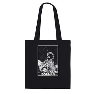 May include: Black tote bag featuring a white graphic of a scorpion under a crescent moon and stars. The text "of SCORPIUS" is below the graphic. The bag has two black handles.