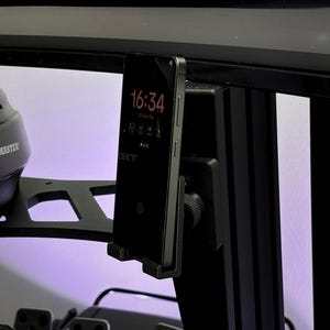 Pode incluir: Um smartphone preto está montado em um suporte preto, exibindo a hora 16:34. O telefone faz parte de uma configuração de simulador de corrida, com um volante e pedais Thrustmaster visíveis. O fundo tem um tom roxo.