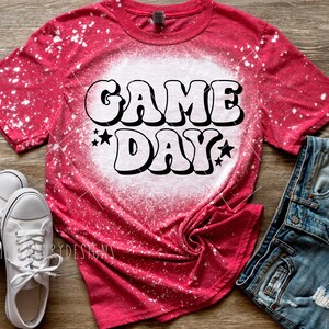 Op de afbeelding: Rood T-shirt met witte bleekmiddelspatten en een witte afbeelding met de tekst "GAME DAY" in een retro-lettertype met sterren.
