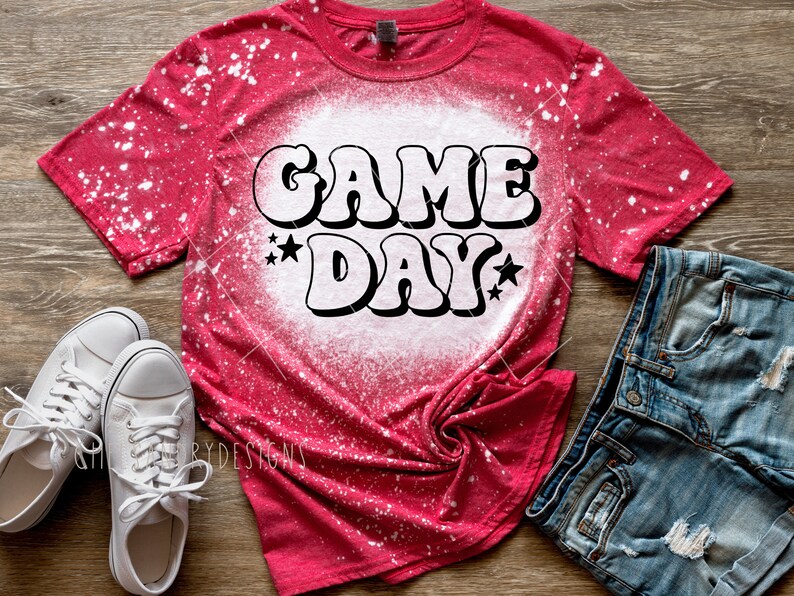 Op de afbeelding: Rood T-shirt met witte bleekmiddelspatten en een witte afbeelding met de tekst "GAME DAY" in een retro-lettertype met sterren.