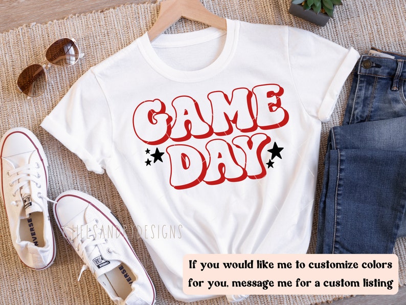 Op de afbeelding: Wit T-shirt met rode en zwarte tekst die "GAME DAY" zegt in een retro lettertype. Het shirt ligt op een bruine ondergrond met een paar witte sneakers, een blauwe jeans en een zonnebril.